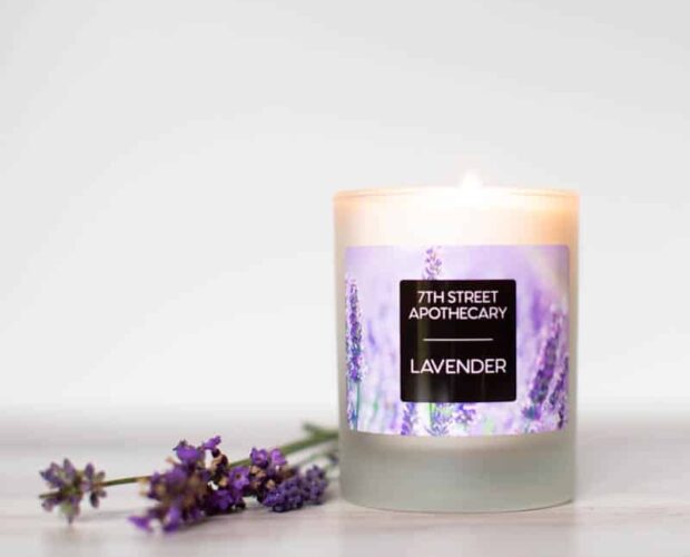 Custom Lavender Label for Homemade and Small Batch Candles. Shows a photo of a candle and lavender flowers. The candle has a label with lavender flowers in the background, with a centered black box with white text reading "7th Street Apothecary - Lavender"